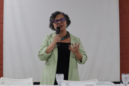 Imagem: Foto da vice-reitora Diana Azevedo em pé segurando um microfone e usando uma blusa preta e um blazer verde claro
