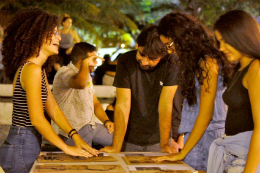 Imagem: Estudantes e um professor em volta de uma mesa, apreciando alguns itens de design em exposição (Foto: Divulgação)