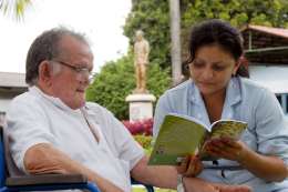 Imagem: Cuidadora lê um livro para idoso sentado em uma cadeira