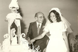 Imagem: Foto de um casal d enoivos no dia do casamento. Homem olha para mulher enquanto ela sorta o bolo de casamento.