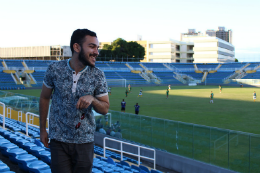 Imagem: Foto de estudante de Jornalismo na arquibancada do Estádio Presidente Vargas e, ao fundo, jogadores no campo 
