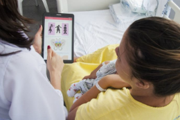 Imagem: Foto de duas mulheres vistas de costas: uma, vestida de jaleco, mostra a outra (paciente) o aplicativo num tablet