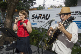 Imagem: Foto de servidor com violão ao lado de servidora cantando no palco do 1° Sarau FazerArte