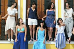 Imagem: Foto do Grupo Damas Cortejam. As integrantes estão encostadas numa fachada com janelas. Alguma estão de pé nas janelas e outras no chão.