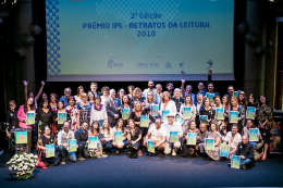 Pessoas com certificados de prêmio reunidas em um palco, sentadas e em pé. Ao fundo, painel escrito 3ª Edição Prêmio IPL - Retratos da Leitura 2018