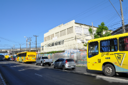 Imagem: Foto de rua onde há uma escola e ônibus e carros circulando no entorno