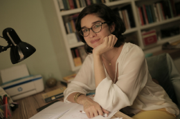 Imagem: Foto de Juliana Diniz apoiada numa escrivaninha onde se vê papéis e uma luminária