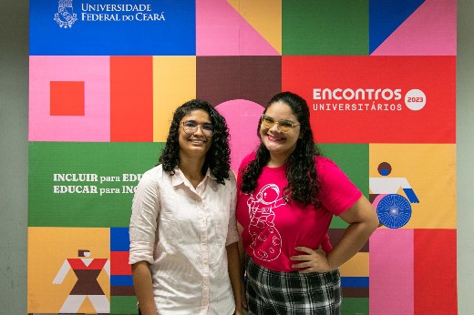 Thalita Batista e Sarah Soares, alunas do Campus de Quixadá, lado a lado, em pé; por trás há um banner do evento (Foto: Viktor Braga/UFC)