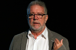 Imagem: foto de um homem branco, de barba e óculos de grau gesticulando como se estivesse falando para um público