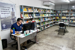 Imagem do interior da livraria, com um funcionário à esquerda, sentado em um birô e acessando um computador. Ao fundo, estantes com seções cheias de livros