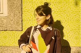 Imagem: Foto da  designer Carla Colares encostada num muro de um prédio e segurando uma bolsa