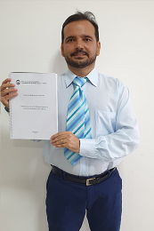 Imagem: Foto do servidor Fábio Luiz segurando sua tese de doutorado nas mãos. Ele veste calça social, camisa social e gravata