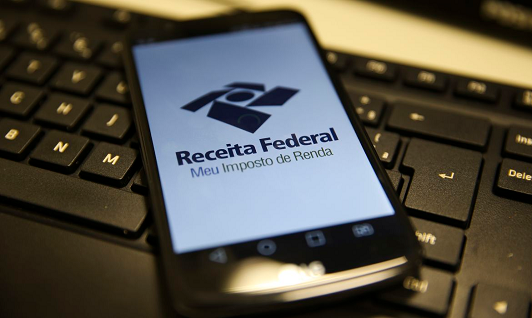 Imagem: foto focando um teclado de computador e, em cima dele, um telefone celular que mostra a tela a marca da Receita Federal