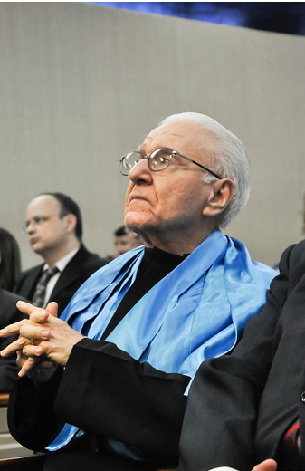 Imagem: Homem idoso, com cabelos brancos, sentado em auditório e vestido com roupa preta e capa azul