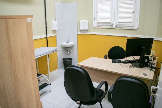 Imagem: ambiente de consultório médico com mesa e computador, três cadeiras pretas e armário.