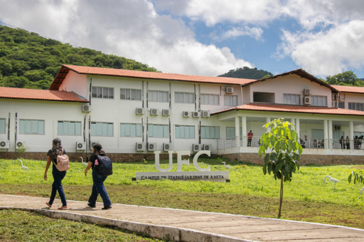 Imagem: Foto da fachada do campus de Itapajé com duas estudantes caminhando em uma calçada transversal. Ao fundo, o verde da paisagem sertaneja