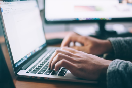 Imagem: Mãos de uma pessoa digitando no teclado de um notebook; ao fundo há um computador de mesa (Foto: Divulgação)