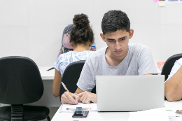 Imagem: Estudante sentado em uma cadeira, com uma pequena mesa a sua frente; ele está olhando para um notebook que há sobre a mesa e fazendo anotações em um caderno; há uma estudante ao fundo, de costas (Foto: Viktor Braga/UFC)