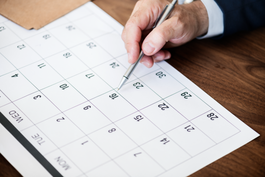 Imagem: calendário de fundo branco e mão segurando uma caneta aponta para uma data