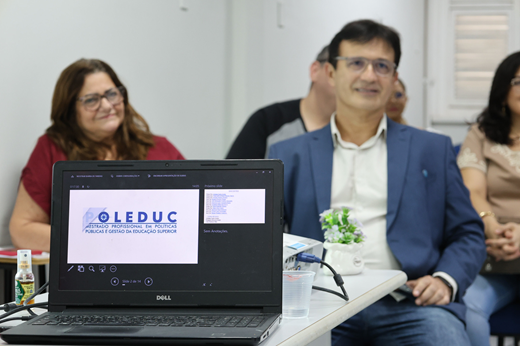Imagem: Em primeiro plano, um notebook aberto exibe apresentação com a logomarca do Poleduc; atrás dele, a pró-reitora Marilene Soares e o reitor Custódio Almeida assistem à fala da coordenadora em exercício