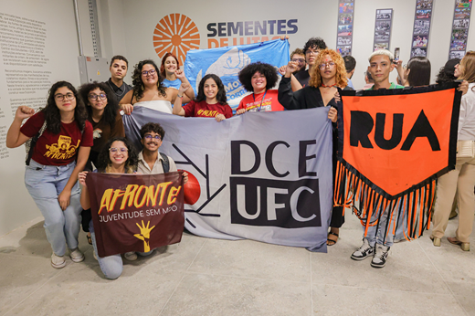 Estudantes posam em frente ao letreiro da exposição Sementes de Lutas com bandeiras do DCE-UFC e de grupos e entidades do movimento estudantil