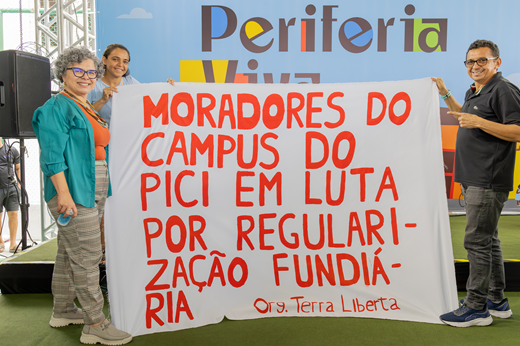 Imagem: Vice-reitora Diana Azevedo posa com uma mulher e um homem, representantes das famílias residentes no Campus do Pici. Eles seguram uma faixa com os dizeres "Moradores do Campus do Pici em luta por regularização fundiária".