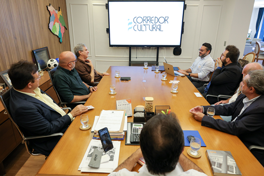 Imagem: Participantes da reunião sentados à mesa no Gabinete da Prefeitura de Fortaleza. No telão, está projetada uma apresentação com o texto "Corredor Cultural"