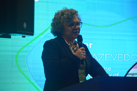 Imagem: Professora Diana Azevedo fala ao microfone no palco; ela traja blazer preto. Ao fundo, há um telão azul.