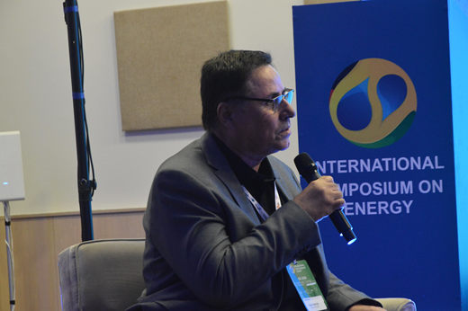 Imagem: Em plano fechado, o professor Enio Pontes fala ao microfone. Ele traja terno cinza. Ao fundo, o telão azul.