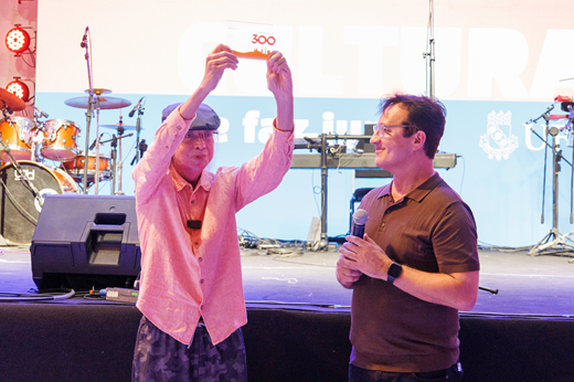 Imagem: Na frente do palco, o cantor Ednardo segura acima da cabeça o troféu Fortaleza 300 anos; ao lado dele, o reitor Custódio Almeida segura o microfone