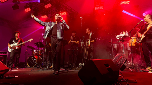 Imagem: Com sua banda, o cantor Marcos Lessa se apresenta no palco como atração se fechamento do evento; a iluminação é predominantemente vermelha