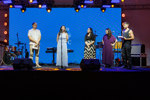 Imagem: No palco, os diretores da Editora da UFC, Cavalcante Júnior e Juliana Diniz, falam ao microfone no lançamento oficial da coletânea Fortaleza Amada; ao lado deles, Tércia Montenegro, Natércia Pontes e Luciano Brito representam os autores da obra.