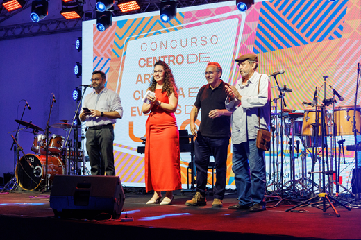 Imagem: No palco, quatro membros da comissão organizadora (uma mulher e três homens) apresentam os termos do concurso de projetos do novo centro cultural da UFC