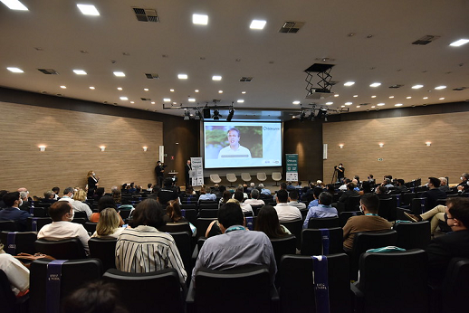 Imagem mostra pessoas sentadas assistindo a telão no qual aparece o governador Camilo Santana