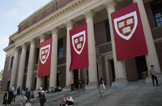 Imagem: fachada da Universidade de Harvard, com paredes vermelhas, colunas brancas, bandeiras penduradas e pessoas sentadas na escadaria localizada a frente