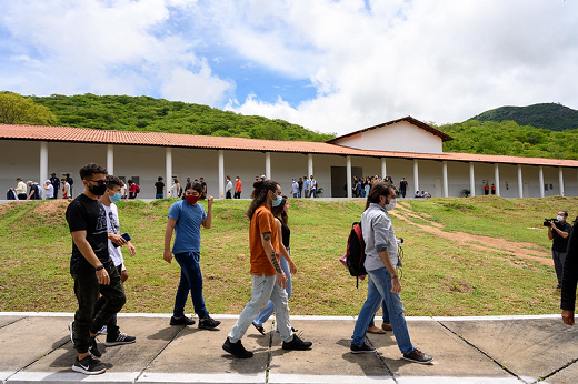 La imagen muestra a los estudiantes caminando en una rampa de acceso a uno de los edificios del campus, que aparece en la parte superior al fondo con varias personas presentes en él.