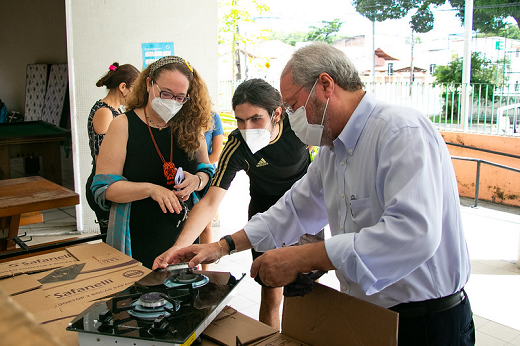 Imagem mostra a professora Geovana Cartaxo à esquerda, o estudante João Vitor ao centro e o reitor Cândido Albuquerque à direita manuseando um dos novos fogões entregues