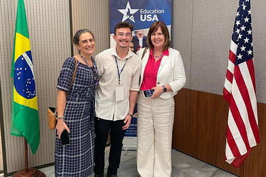A imagem mostra três pessoas sorrindo, posando para uma foto em ambiente interno. Elas estão de pé, lado a lado, com duas bandeiras ao fundo — à esquerda, a bandeira do Brasil; à direita, a bandeira dos Estados Unidos. Entre elas, há um banner com a logomarca do programa “EducationUSA”.  À esquerda, está uma mulher de pele clara e cabelos grisalhos presos. Ela veste um vestido longo com estampa em xadrez azul e branco, segura um celular na mão direita e uma bolsa caramelo no ombro esquerdo.  Ao centro, há um homem jovem de pele clara, cabelo castanho curto e barba rala. Ele está sorrindo e veste uma camisa branca de manga curta, calça preta e crachá pendurado no pescoço.  À direita, há uma mulher de pele clara e cabelo castanho claro, liso e na altura dos ombros. Ela veste uma blusa rosa, blazer branco e calça branca. Também está sorrindo e segura um celular com a mão esquerda.