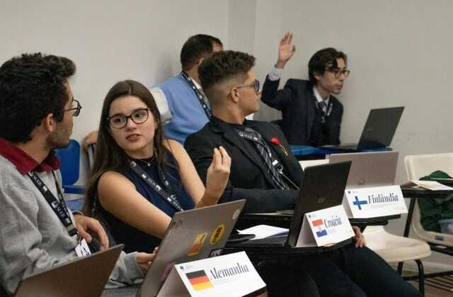 imagem: um grupo de jovens participa de uma simulação diplomática em sala de aula. Eles estão sentados em carteiras com notebooks abertos, cada um identificado com uma placa que traz o nome e a bandeira de um país, como Alemanha, Croácia e Finlândia. Uma jovem de óculos, com vestido azul escuro, conversa com um colega ao lado. Outro rapaz, de terno preto, olha para trás, enquanto, mais ao fundo, um participante de paletó azul ergue a mão para pedir a palavra. O ambiente é de debate e discussão, lembrando uma reunião de organismos internacionais.