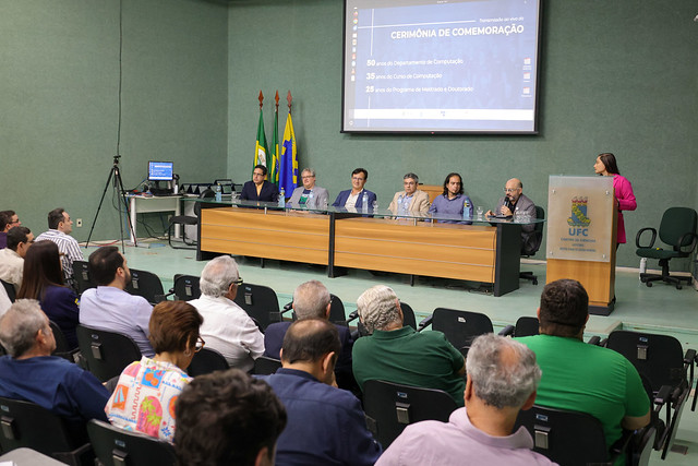 imagem: Auditório do Centro de Ciências da Universidade Federal do Ceará, com público sentado assistindo a uma mesa de autoridades composta por sete pessoas. Elas estão posicionadas atrás de uma longa bancada de madeira, em frente a um telão que exibe o título “Cerimônia de Comemoração: 50 anos do Departamento de Computação, 35 anos do Curso de Computação e 25 anos do Programa de Mestrado e Doutorado”. À direita, uma mulher de blazer rosa fala ao microfone em um púlpito com o brasão da UFC. Ao fundo, há bandeiras do Brasil, do Ceará e da Universidade.