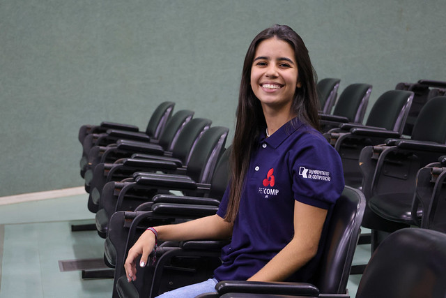 imagem: Mulher jovem, de cabelos longos e lisos, sorri sentada em uma das cadeiras do auditório do Centro de Ciências da Universidade Federal do Ceará. Ela usa camisa azul-marinho com o logotipo do Programa de Educação Tutorial de Computação (PETComp) e do Departamento de Computação da UFC bordados na manga e no peito. Ao fundo, aparecem fileiras de cadeiras vazias.