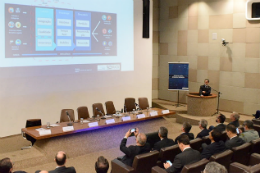 imagem: foto de auditório com pessoas sentadas e um homem no palco, apresentando slides projetados numa tela