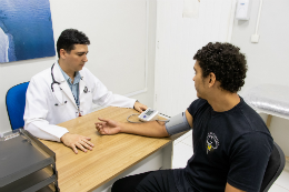Imagem: Médico medindo pressão arterial de um homem, ambos sentados próximos a uma mesa (Foto: Viktor Braga/UFC)