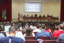 Imagem: foto a partir do fundo do auditório, onde se pod ver o público sentado e o palco, com mesa de autoridades e reitor falando ao púlpito