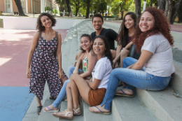 Imagem: Foto de cinco estudantes (1 homem e 4 mulheres) sentados na escada do palco na concha acústica e de uma estudante em pé