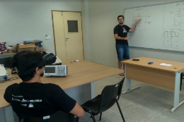 Imagem: Foto de um estudante em sala de aula com óculos adaptado observando a explicação de um professor na lousa