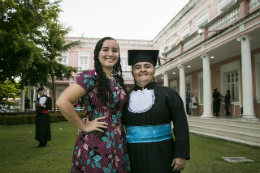 Imagem: Foto de uma senhora vestida de beca ao lado da filha, uma jovem de cabelos longos e cacheados usando vestido estampado