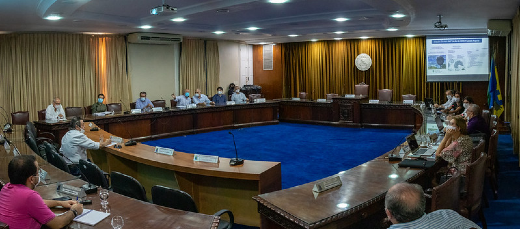 Foto panorâmica da Sala do Conselho Universitário da UFC, com os participantes tomando assento. São cinco mesas de madeira de borda circular, posicionadas em um quadrado. O piso é um carpete azul, e no fundo há uma cortina com o brasão da UFC esculpido em gesso.