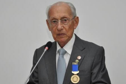 Foto do Prof. Dilson Menezes utilizando um terno e uma gravata de cor cinza, uma medalha posta na lapela discursando