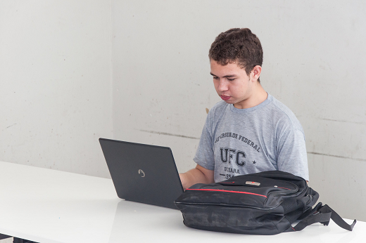 Imagem: Estudante sentando em una cadeira, com um notebook à sua frente, sobre uma mesa (Foto: Viktor Braga/UFC)